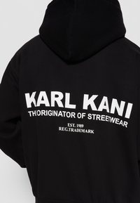 Sweat à capuche noir avec texte blanc "KARL KANI THE ORIGINATOR OF STREETWEAR EST. 1989 REG. TRADEMARK" dans le dos.