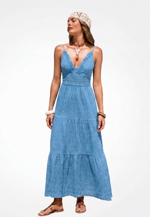 Donna con un vestito lungo blu senza maniche con dettagli in pizzo, una fascia per capelli all'uncinetto, collane sovrapposte, braccialetti e sandali bassi marroni.