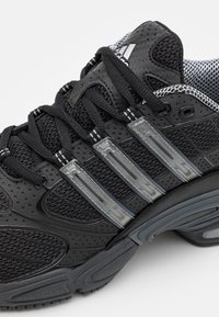 Svart atletisk sko med ovandel av mesh med perforerad textur, tre grå ränder och en böjd gummisula. Snören är svarta.