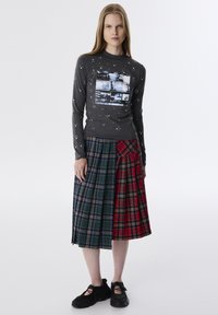 Grijze langere mouwtrui met een fotoprint en witte stipaccenten, gecombineerd met een geruite rok in groene en rode tinten met plooien. Zwarte sneakers.