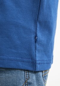 Una camiseta de algodón azul presenta una tela texturada con una pequeña etiqueta de la marca en el dobladillo, combinada con jeans de mezclilla que muestran detalles de costura visibles.
