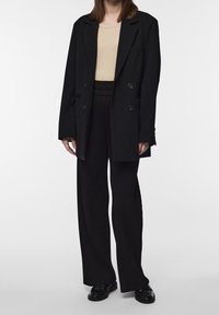 Blazer noir à double boutonnage sur un haut beige côtelé, associé à un pantalon large côtelé noir. Chaussures en cuir verni noir.