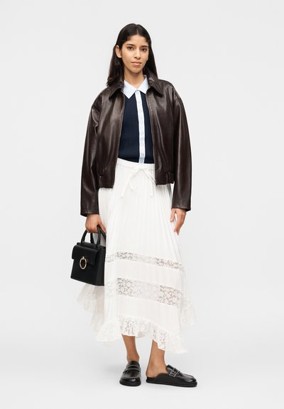 Γυναίκα που φοράει σκούρο καφέ δερμάτινο μπουφάν, μπλε ναυτικό πουλόβερ με κουμπιά, λευκή δαντελένια φούστα, μαύρα μοκασίνια και κρατά μια μαύρη τσάντα χειρός.