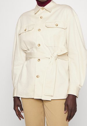 Veste crème boutonnée avec poches poitrine et ceinture nouée à la taille, portée sur un col roulé bordeaux et un pantalon beige.