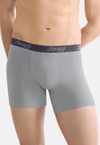 Grijze Baumwoll-Boxer mit enger Passform und elastischem Bund, der das "sloggi"-Logo in Weiß und Rot zeigt. Glatte Textur, bis zur Mitte des Oberschenkels.