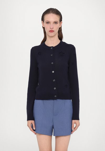 Marineblå strik cardigan med knaplukning, ribbet kant og fem dekorative stjerneapplikationer på forsiden. Kombination med lyseblå shorts.