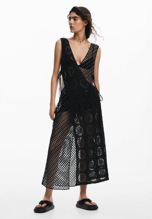 Woman wearing a sleeveless black crochet maxi dress over black underwear, paired with black platform sandals, standing against a white background.