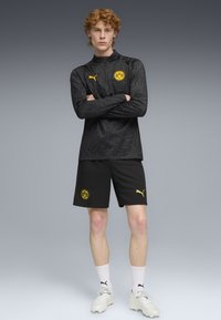 Top deportivo negro con cremallera y un patrón sutil, logo de Puma amarillo y escudo de Dortmund, combinado con pantalones cortos negros que presentan el mismo logo.