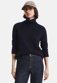 Pulover de tip guler înalt de culoare bleumarin, dintr-un material moale, cu manșete cu nervuri, purtat împreună cu blugi de culoare închisă. Capac de culoare gri deschis cu vizieră maro și logo.
