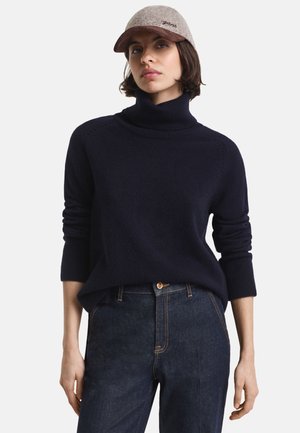 Marineblå turtleneck-genser i mykt stoff med ribbestrikkede mansjetter, sammen med mørke denimjeans. Capen er lysegrå med brunskjerm og logo.