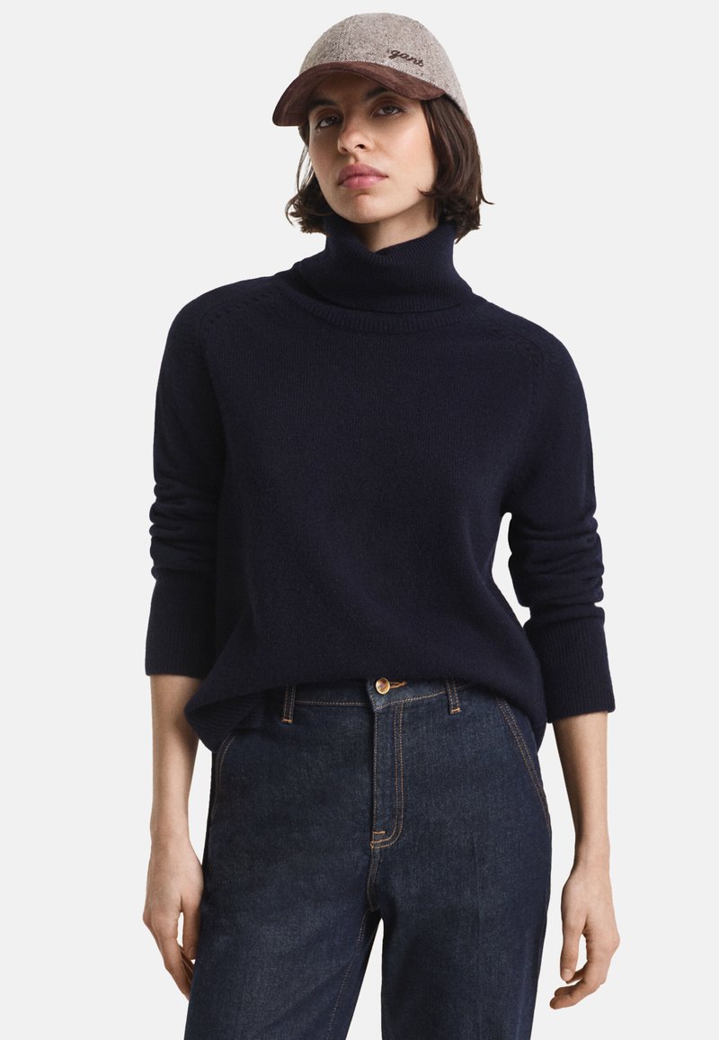 Pull à col roulé marine en tissu doux avec des poignets côtelés, associé à un jean en denim foncé. La casquette est gris clair avec une visière marron et un logo.