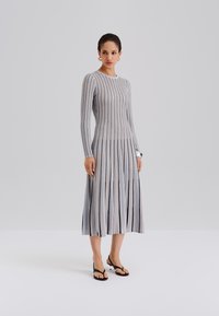 Dzianinowa sukienka z długimi rękawami, ozdobiona pionowymi pasami w odcieniach szarości, niebieskiego i czerni. Rozkloszowany krój i długość do kostek; noszona z czarnymi japonkami.