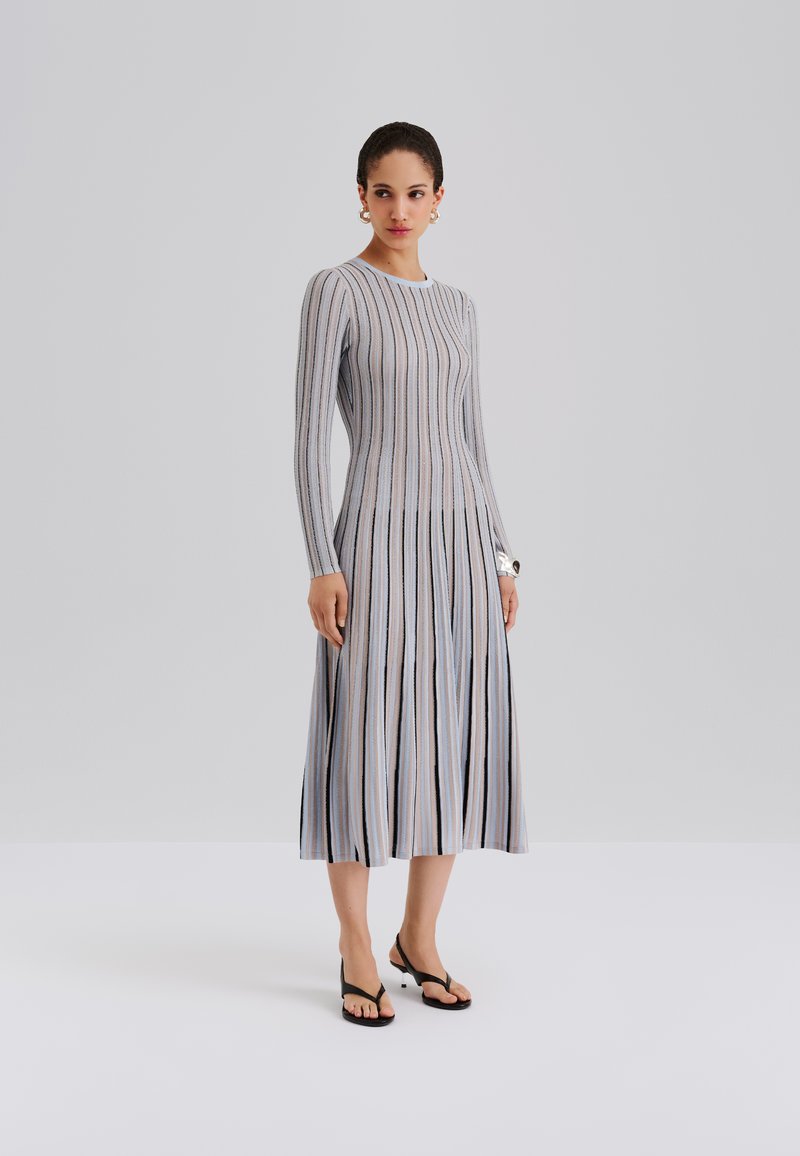 Dzianinowa sukienka z długimi rękawami, ozdobiona pionowymi pasami w odcieniach szarości, niebieskiego i czerni. Rozkloszowany krój i długość do kostek; noszona z czarnymi japonkami.