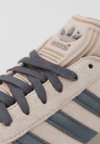 Tenisky Adidas s béžovou semišovou úpravou, černými tkaničkami a černými podpisovými třemi pruhy na boku, detailní záběr na jazyk a tkaničky.