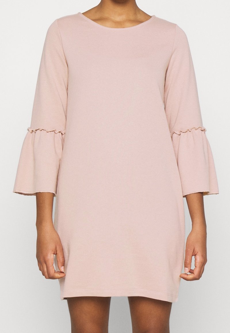 Femme portant une robe rose pâle, longueur genou, avec des manches cloche trois-quarts ornées de coutures volantées aux coudes.