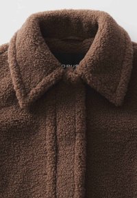 Veste en polaire marron avec une surface douce et texturée, dotée d'un col rabattu et d'une patte de boutonnage droite. Étiquette de la marque visible à l'intérieur.