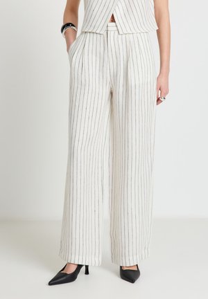 Femme portant un pantalon blanc à jambes larges avec de fines rayures verticales noires, associé à des escarpins noirs à bouts pointus et un haut court à rayures.