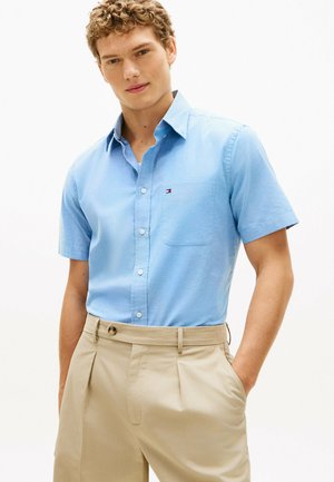 Młody mężczyzna ubrany w jasnoniebieską, krótkorękawową koszulę zapinaną na guziki oraz beżowe plisowane spodnie, ze schowanymi w kieszeniach rękami, stojący na jednolitym tle.