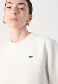 Felpa bianca con finitura testurizzata, caratterizzata da un piccolo logo di coccodrillo verde sul petto. Collo rotondo e spalle strutturate.