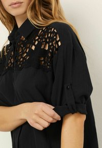 Prim-plan al unei persoane care își suflecă mâneca unei cămăși negre cu detalii din dantelă în zona superioară a pieptului și a umărului.