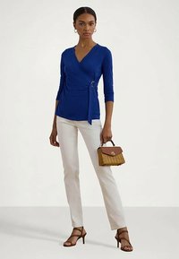 Γυναίκα φορώντας μια βασιλική μπλε μπλούζα τύπου wrap, λευκά παντελόνια, καφέ ψηλοτάκουνα πέδιλα και κρατώντας μια μικρή πλεκτή τσάντα με καφέ καπάκι.