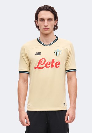 Un homme porte un maillot de football de l’Atalanta jaune pâle, orné de bordures noires et bleues et du nom du sponsor Lete en rouge, sur un fond uni.