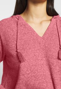 Roze, gemêleerde V-hals top met een zachte textuur, voorzien van twee franjeaccenten aan de trekkoorden van de capuchon.
