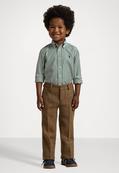 Χαρούμενο παιδί με σγουρά μαλλιά φοράει πράσινη και λευκή ριγέ πουκάμισο, καφέ υφασμάτινα παντελόνια και ναυτικό μπλε αθλητικά παπούτσια, στέκεται σε λευκό δάπεδο.