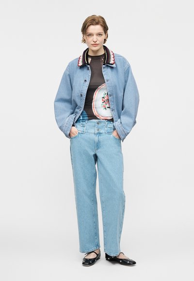 Άτομο που φοράει ανοιχτό μπλε τζιν μπουφάν με σχέδιο στο γιακά, μαύρο γραφικό πουκάμισο, ανοιχτό μπλε ψηλόμεσο τζιν και μαύρες μπαλαρίνες με ασημένιες καρφίτσες.