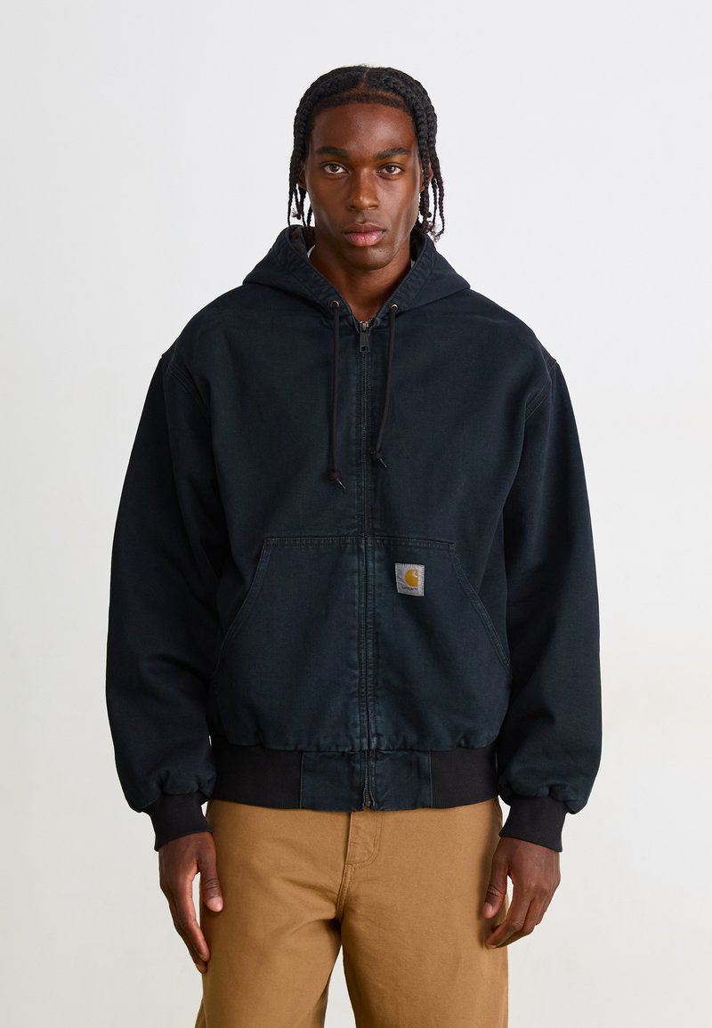 Man with braided hair wearing a black zip-up hoodie and tan pants standing against a plain white background.