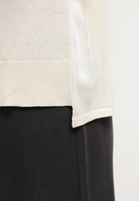 Krämfärgad stickad topp med ribbat mönster och kontrasterande slät tyg vid fållen, lager-på-lager över ett svart plagg.