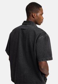 Camicia nera in denim a maniche corte con vestibilità rilassata, caratterizzata da un colletto, una spalla dietro e un piccolo etichetta vicino al collo.