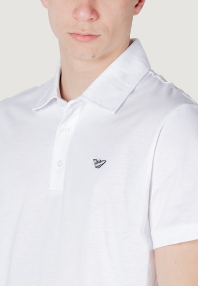 Polo shirt blanc en tissu doux, doté d'un col classique, d'une patte de boutonnage à trois boutons et d'un petit logo noir brodé sur la poitrine.