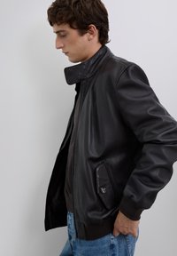 Junger Mann im Profil, der eine schwarze Lederjacke, einen dunklen Pullover und blaue Jeans trägt, und vor einem einfarbigen hellgrauen Hintergrund steht.