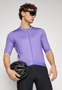 Fialové cyklistické tričko s krátkym rukávom a predným zipsom, v kombinácii s čiernymi cyklistickými šortkami a čiernou prilbou, držané v ruke.