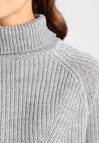 Maglione lavorato a maglia grigio chiaro con collo alto a coste e un motivo texturizzato, caratterizzato da una vestibilità rilassata e dettagli di cucitura visibili.