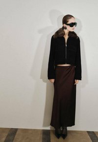 Černá zkrácená bunda s kožešinovým límcem, doplněná dlouhou hnědou sukní. Modelka má velké černé sluneční brýle a špičaté černé lodičky.