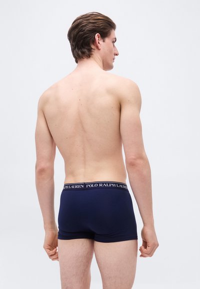 Νεαρός άνδρας όρθιος με την πλάτη να κοιτά μπροστά, φορώντας εσώρουχα τύπου boxer Polo Ralph Lauren σε ναυτικό μπλε χρώμα, μπροστά σε απλό φωτεινό φόντο.