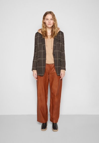 Blazer xadrez castanho com lapelas de bico, usado sobre um hoodie bege, combinado com calças de couro castanhas de perna larga e botas de plataforma pretas.