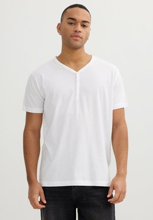 Hombre joven de pie con una camisa Henley blanca de manga corta y pantalones negros, sobre un fondo claro y liso.