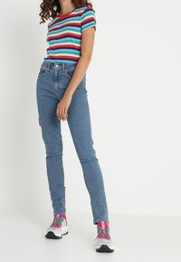Jeans ajustados de mezclilla azul combinados con una camiseta de rayas multicolores. La camiseta tiene mangas cortas y un escote redondeado. El calzado notable incluye zapatillas deportivas con una base blanca y acentos en rosa intenso.
