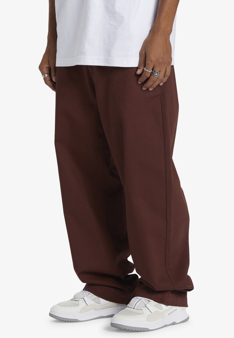 Personne portant un pantalon ample marron foncé, des baskets blanches et une chemise blanche, avec plusieurs bagues argentées visibles aux doigts.