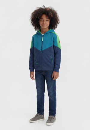 Dieťa stojace, oblečené v modro-zelenú mikinu na zips, tmavomodré džínsy a sivé tenisky, na jednoduchom bielom pozadí.