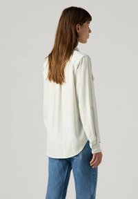 Langärmlige, hellbeige Bluse aus weichem Stoff, mit lockerer Passform, Knopfmanschetten und geradem Saum. Getragen mit blauen Jeans.