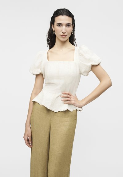 Γυναίκα με σκούρα μαλλιά φοράει λευκή μπλούζα με φουσκωτά μανίκια και τετράγωνο ντεκολτέ, καθώς και ανοιχτό καφέ παντελόνι με φαρδιά πόδια, ποζάροντας με το ένα χέρι στη μέση.