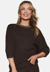 Brauner Strickpullover mit kurzen Ärmeln, der einen wellenförmigen Ausschnitt mit goldenen Verzierungen aufweist. Strukturierter, gemütlicher Stoff.