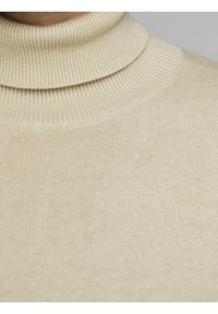 Beige gestrickter Rollkragenpullover mit einem dicken, gerippten Kragen und glatter Textur, mit einer Nahaufnahme des Stoffmusters.