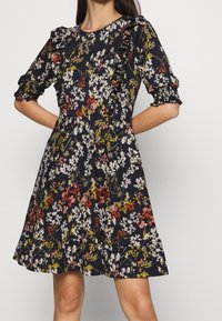 Femme portant une robe fleurie noire avec des volants sur la poitrine et les manches, ornée de fleurs roses, blanches et vertes, et dotée d'une jupe évasée.