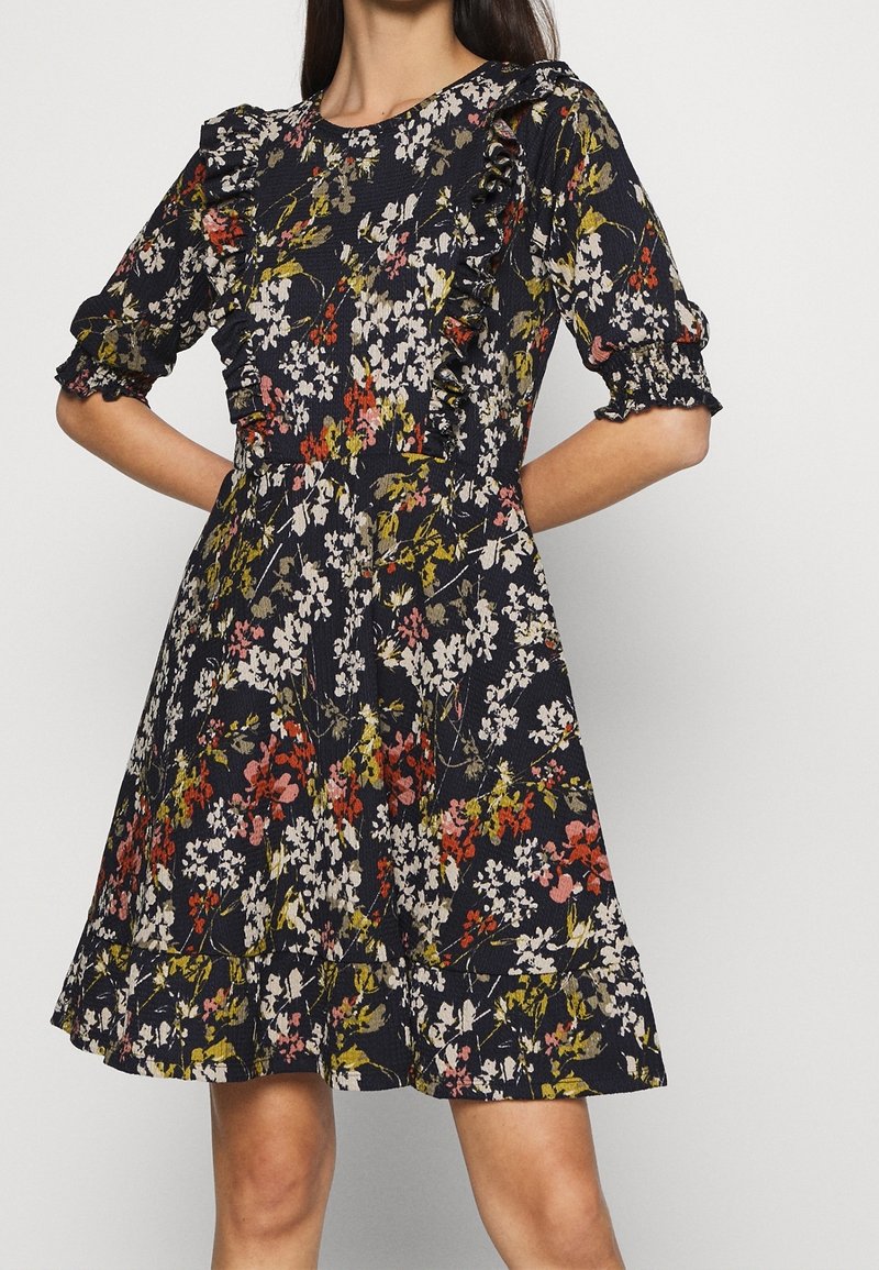 Femme portant une robe fleurie noire avec des volants sur la poitrine et les manches, ornée de fleurs roses, blanches et vertes, et dotée d'une jupe évasée.