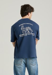 Mørkeblå kortærmet t-shirt lavet af blødt stof med en løs pasform. Har hvide trykte grafik på ryggen, inklusive tekst og figurer.
